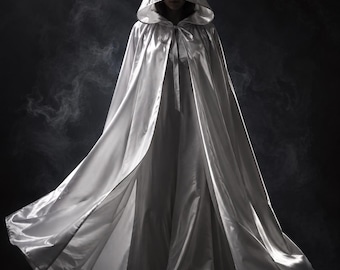 May include: A floor-length white satin cloak featuring a deep hood and a ribbon tie closure at the neck. The garment is constructed from a smooth, reflective fabric that drapes fully to the floor, creating a voluminous silhouette with wide folds.