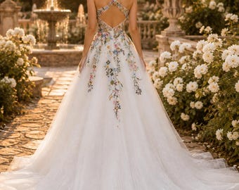 May include: A floor-length white gown featuring a sleeveless bodice with a crisscross back design and a full skirt. The dress is adorned with colorful floral embroidery that extends from the bodice down the center of the skirt.