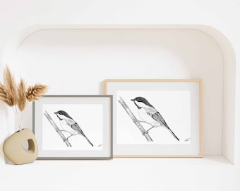 May include: Two framed art prints featuring a black and white ink illustration of a chickadee perched on a thin branch. The prints are displayed in a gray frame and a light wood frame, accompanied by a beige ceramic vase with dried grass stems.