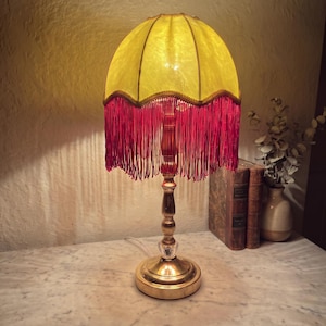 May include: A table lamp with a metallic gold base and a translucent yellow lampshade. The shade features a scalloped bottom edge adorned with long, deep red fringe. The lamp stand has a slender, tiered design with a clear crystal accent.