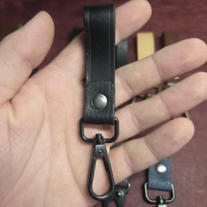 May include: A black leather key fob strap featuring a silver-toned metal snap button closure and a matte black swivel lobster clasp. The strap is folded over to create a loop, with the metal hardware securely attached to the bottom of the leather.