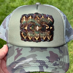 May include: An olive green trucker hat featuring a camouflage print on the brim and a rectangular patch on the front panel. The patch is composed of layered, iridescent feathers in shades of brown, black, and metallic green with pointed tips.