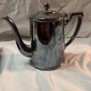 May include: A polished silver-toned metal coffee pot featuring a cylindrical body, a curved spout, and a hinged lid topped with a small spherical finial. The handle is angular and attached to the side, with decorative grooved bands at the top and base.