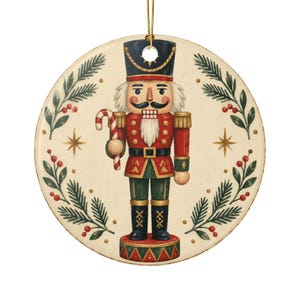 May include: A round ceramic ornament featuring a central illustration of a nutcracker soldier in a red, green, and black uniform holding a red and white candy cane. The design is framed by green pine branches with red berries and gold stars.