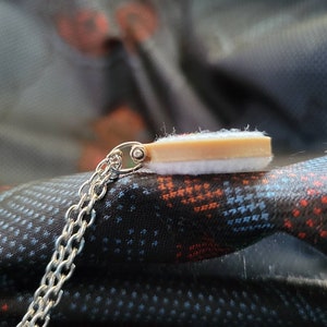 May include: A silver chain necklace with a small pendant. The pendant has a rectangular shape with a light brown top and a white bottom. The necklace is displayed on a patterned fabric.
