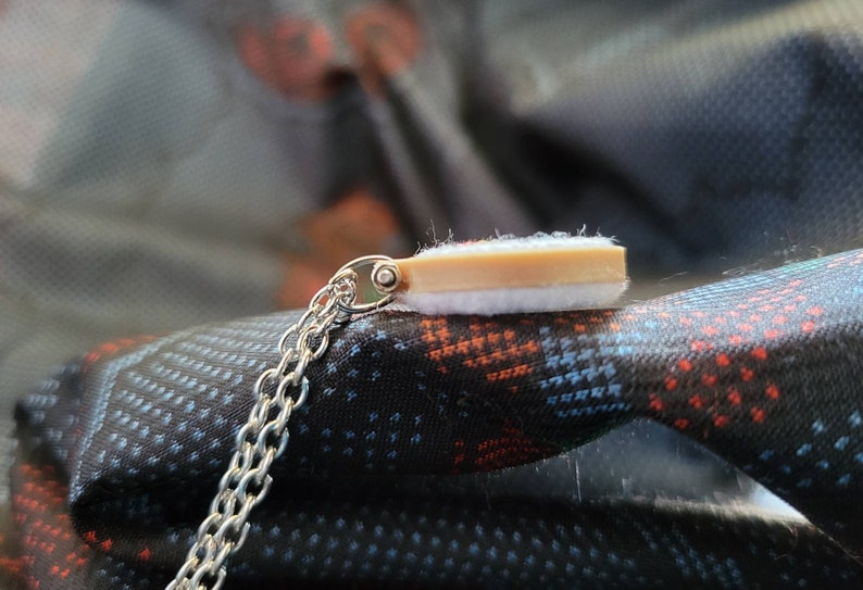 May include: A silver chain necklace with a small pendant. The pendant has a rectangular shape with a light brown top and a white bottom. The necklace is displayed on a patterned fabric.
