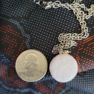 May include: A silver chain necklace with a small, round, white felt pendant. A U.S. quarter coin is next to the pendant for size comparison. The background is a dark fabric with a pattern of small white dots and red and blue shapes.