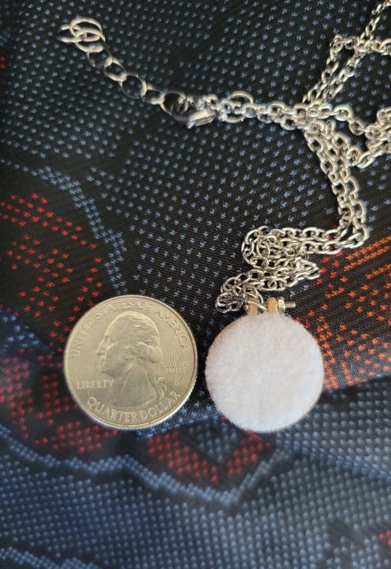 May include: A silver chain necklace with a small, round, white felt pendant. A U.S. quarter coin is next to the pendant for size comparison. The background is a dark fabric with a pattern of small white dots and red and blue shapes.