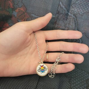 May include: A silver necklace with a small, round pendant featuring a colorful embroidered bee design. The bee has rainbow-colored wings and a black outline. The pendant is set in a small wooden hoop.