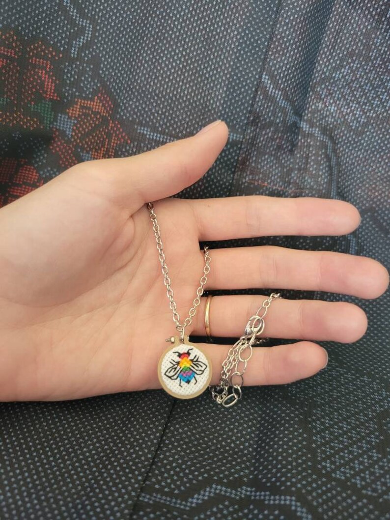 May include: A silver necklace with a small, round pendant featuring a colorful embroidered bee design. The bee has rainbow-colored wings and a black outline. The pendant is set in a small wooden hoop.