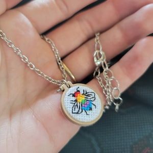 May include: Silver-toned necklace featuring a small, round pendant with a cross-stitch design of a bee in rainbow colors. The bee is stitched on white fabric, set within a miniature wooden embroidery hoop. The chain has a lobster clasp.