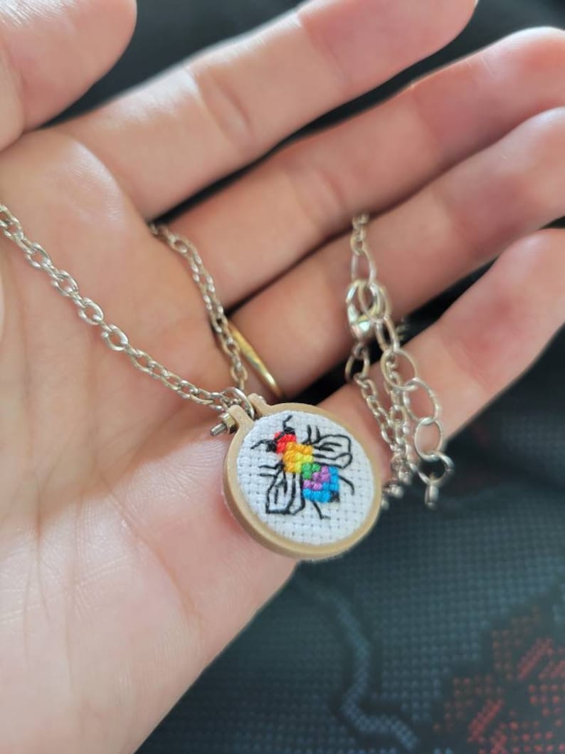 May include: Silver-toned necklace featuring a small, round pendant with a cross-stitch design of a bee in rainbow colors. The bee is stitched on white fabric, set within a miniature wooden embroidery hoop. The chain has a lobster clasp.