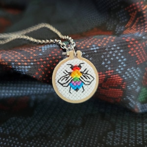 May include: A silver chain necklace with a small, round pendant. The pendant features a cross-stitch design of a bee with rainbow-colored wings and a black outline, set in a wooden embroidery hoop.