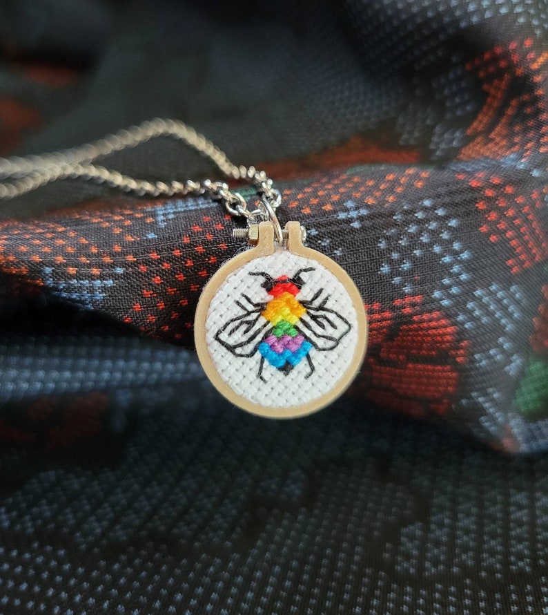 May include: A silver chain necklace with a small, round pendant. The pendant features a cross-stitch design of a bee with rainbow-colored wings and a black outline, set in a wooden embroidery hoop.