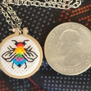 May include: A small, round pendant necklace featuring a cross-stitched bee with rainbow-colored wings. The bee is set in a miniature embroidery hoop. The necklace chain is silver-colored. A U.S. quarter coin is next to the pendant for size comparison.