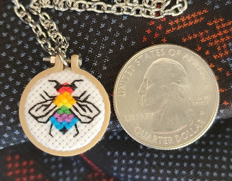 May include: A small, round pendant necklace featuring a cross-stitched bee with rainbow-colored wings. The bee is set in a miniature embroidery hoop. The necklace chain is silver-colored. A U.S. quarter coin is next to the pendant for size comparison.
