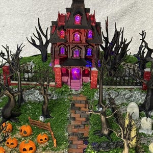 May include: A miniature haunted house model with a dark roof and red brick walls, illuminated by purple interior lights. The scene includes a brick pathway, bare black trees, a small fence, several orange jack-o'-lanterns, and tombstones.