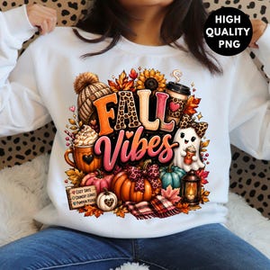 May include: A white sweatshirt featuring a colorful graphic with the text "FALL VIBES" and "COZY DAYS, CRUNCHY LEAVES, PUMPKIN PLEASE". The design includes pumpkins, a ghost, a knit hat, a coffee cup, a lantern, and autumn leaves in orange and brown.
