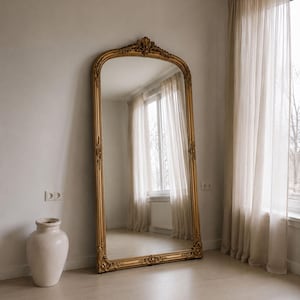 Puede incluir: Un espejo de pie alto con un marco dorado arqueado y ornamentado que presenta intrincados detalles tallados en la parte superior y los lados. El espejo tiene una superficie de vidrio reflectante y está colocado contra una pared clara.