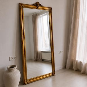 Puede incluir: Un espejo de suelo rectangular y alto con un marco grueso y ornamentado de color dorado, que presenta detalles decorativos en la parte superior central. El espejo está apoyado contra una pared y refleja una habitación con una ventana y cortinas claras.