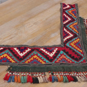 May include: A colorful embroidered fabric trim with a geometric pattern and fringe. The trim is made of a dark red fabric with black, white, blue, green, yellow, and orange embroidery. The fringe is made of green yarn and has small beads.