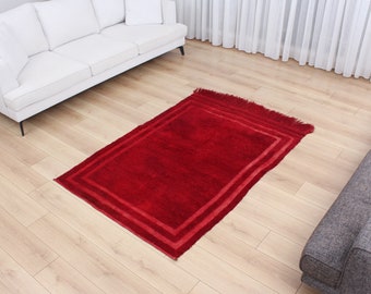 May include: A red rug with a double border in a lighter shade of red. The rug has fringe on two sides and is laid out on a wooden floor.