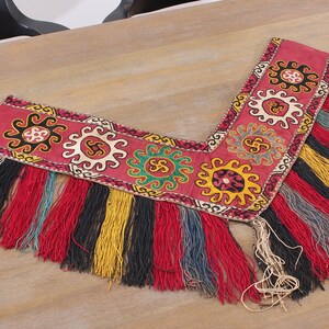 May include: A red, embroidered fabric trim with a fringe of white, red, black, yellow, and blue threads. The trim features a repeating pattern of stylised suns and swirls.