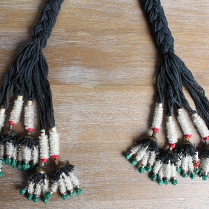 May include: Two black braided cords with white and green beads and black tassels. The cords are tied together at the top and have a red bead at the end of each cord.