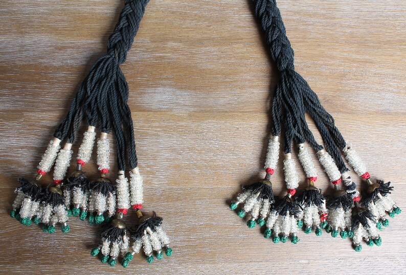 May include: Two black braided cords with white and green beads and black tassels. The cords are tied together at the top and have a red bead at the end of each cord.