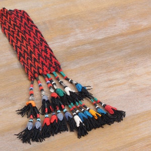 May include: A red and black woven strap with a fringe of colourful tassels and beads. The tassels are made of black yarn and have small beads in various colours, including blue, green, red, and white.