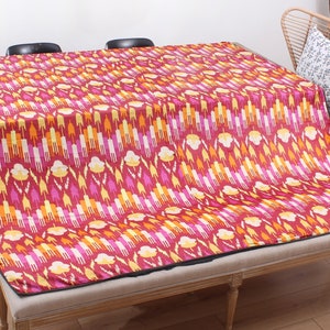 Könnte beinhalten: Eine rote, gelbe und rosafarbene gemusterte Wolldecke mit schwarzem Rand. Die Decke ist gefaltet und über eine Holzbank gelegt.