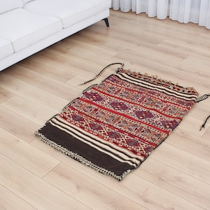 May include: A traditional handwoven rug with a geometric pattern in shades of red, white, and black. The rug has a fringe border and is made of wool.