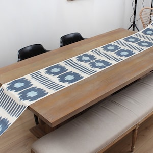 Puede incluir: Un camino de mesa blanco con un patrón ikat azul y blanco. El camino de mesa está colocado sobre una mesa de madera con sillas negras y un banco.