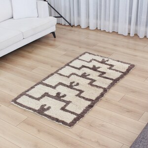 May include: A white and brown geometric patterned rug with a shaggy texture. The rug is laid out on a light brown wood floor.