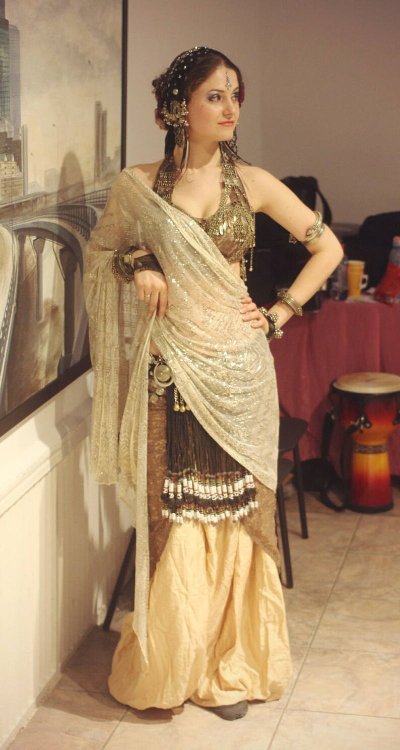 May include: A woman wearing a gold and silver beaded top and a sheer, sparkly, beige sari. She has a bindi on her forehead and a jeweled headpiece.