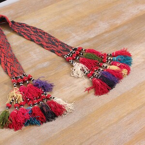 Op de afbeelding: Een geweven textielen riem met een rood-zwart patroon. De riem heeft meerdere kwasten in verschillende kleuren, waaronder rood, paars, blauw en crème. De kwasten zijn met zwarte en witte kralen aan de riem bevestigd. De riem ligt op een lichtbruin houten oppervlak.