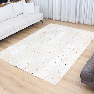 May include: A white area rug with a patchwork design featuring small colorful accents. The rug has a subtle texture and is laid out on a hardwood floor.