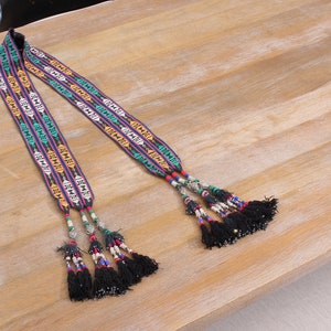 May include: Two long, colourful, woven straps with black tassels and silver beads. The straps have a geometric pattern in shades of blue, green, yellow, and red.