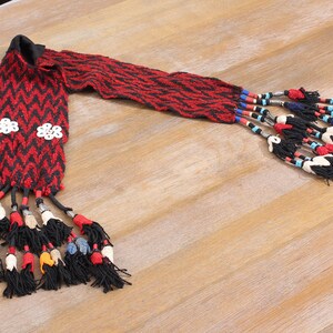 May include: A woven red and black scarf with a chevron pattern. The scarf has white flower-shaped embellishments and black tassels with colorful beads and accents. The scarf is displayed on a light wood surface.