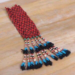 Op de afbeelding: Een geweven textiel met een rood en zwart chevronpatroon. Het textiel heeft meerdere kralenkwasten met zilverkleurige metalen accenten, zwart en blauw garen en kleurrijke kralen. Het item wordt gepresenteerd op een licht houten oppervlak.