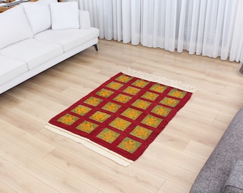 May include: A red and green patterned rug with a white border and fringe. The rug has a geometric design with squares and circles.