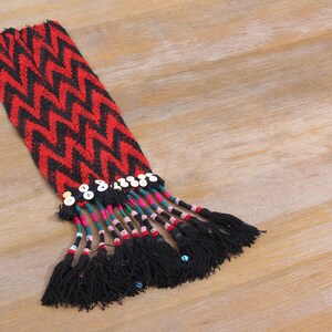 Op de afbeelding: Een rood en zwart geweven stof met een chevron-patroon en een franje van zwarte kwastjes. De franje is versierd met witte knopen en gekleurde kralen.