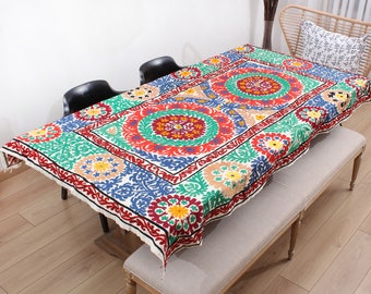 Puede incluir: Un mantel blanco con coloridos patrones florales bordados en tonos rojos, verdes, azules y amarillos. El mantel está puesto sobre una mesa de madera con dos sillas negras y un banco.