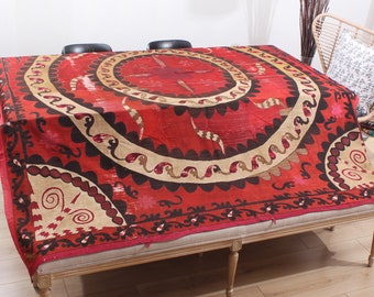 May include: A red and cream colored vintage tablecloth with a circular design featuring intricate embroidery. The tablecloth is laid out on a wooden table with two black chairs.