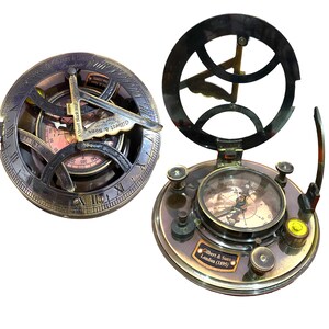 May include: A brass sundial compass with a dark metallic finish. The device features engraved Roman numerals, a central gnomon, and a circular base. A small plaque reads Gilbert & Sons London 1895. The compass includes adjustable knobs and a level.