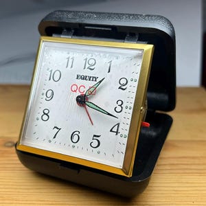 May include: A square Equity travel alarm clock with a gold-tone frame and a white face featuring black Arabic numerals. The clock has black hour and minute hands with green luminous accents, a red second hand, and the text QC printed in red.