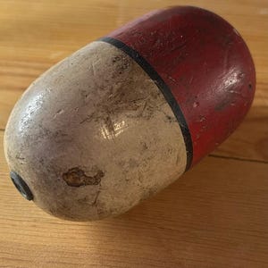 May include: A cylindrical wooden fishing float with a rounded end, featuring a weathered white lower section and a red upper section separated by a thin black band. The surface shows signs of wear, including scratches and small chips in the paint.