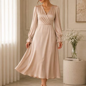 May include: A champagne-colored satin midi dress featuring a V-neckline with a row of small buttons, long sleeves with gathered cuffs, and a wrap-style bodice with pleats at the waist, flowing into a flared skirt that falls below the knees.