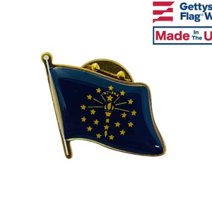 May include: A gold-colored pin with a blue and gold Indiana state flag design. The flag is waving and has a gold star in the center with a torch and a sunburst. The text "Indiana" is written in gold on the flag. The pin is made in the USA.