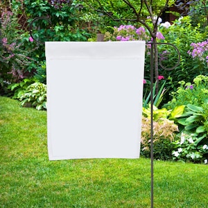 May include: A white blank garden flag is hanging on a black metal flagpole in a garden setting. The flag is surrounded by green grass and colorful flowers.